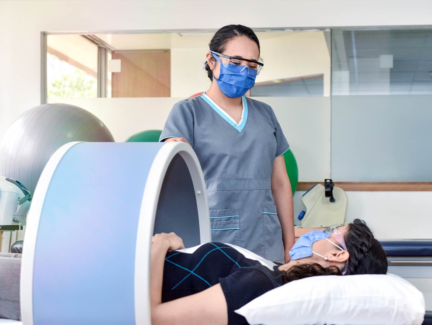 Paciente recostada sobre un equipo de magnetoterapia de color azul con blanco, mientras ve fijamente a la fisioterapeuta